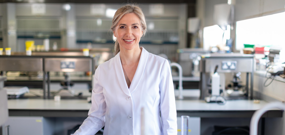 Fabulous pharmacist, Laura Dowling stands in a science lab surrounded by lab equipment- a conical flask, graduated cylinder, flowers ,herbs and mushrooms. She is wearing a bright white, starched lab coat. Her hair is tied back in a loose ponytail 