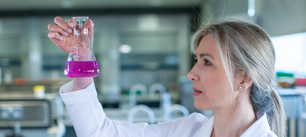 Laura Dowling, fabulous pharmacist, standing in a science lab. She is wearing a white lab coat with blonde hair tied back in a loose ponytail. She is looking closely at and  holding a conical flask with purple liquid inside.